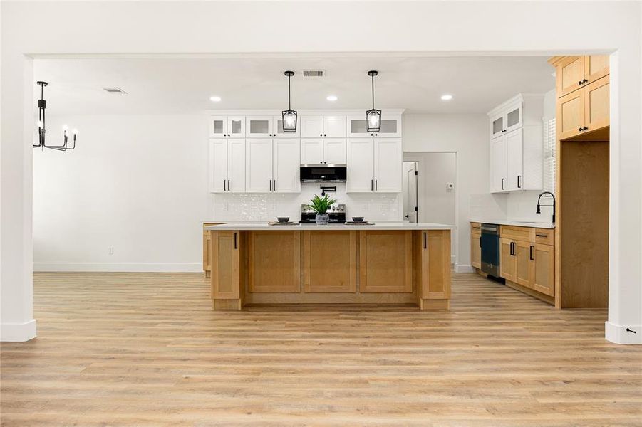 This photo showcases a spacious, modern kitchen with a large island and wooden cabinetry. It features white upper cabinets, stainless steel appliances, and stylish pendant lighting. The open layout extends into a dining area with hardwood flooring, creating an inviting and functional space.