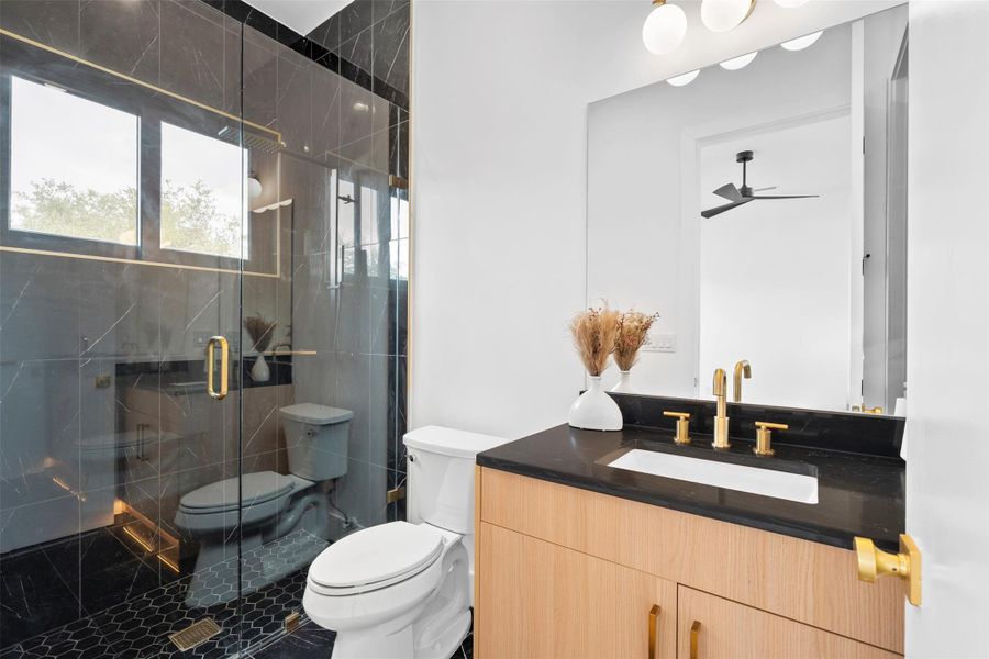 This bathroom features a contemporary vanity with a dark countertop and gold-toned fixtures, a spacious mirror, and a glass-enclosed shower with dark tiled walls and a hex-tiled floor