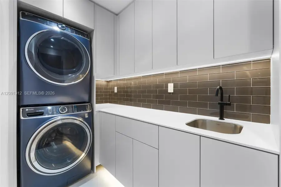 Laundry area featuring stacked front-load appliances, quartz countertops, high-gloss cabinetry, a stainless steel undermount sink with a matte black faucet, and a tiled backsplash Laundry area featuring stacked front-load appliances, quartz countertops, high-gloss cabinetry, a stainless steel undermount sink with a matte black faucet, and a tiled backsplash