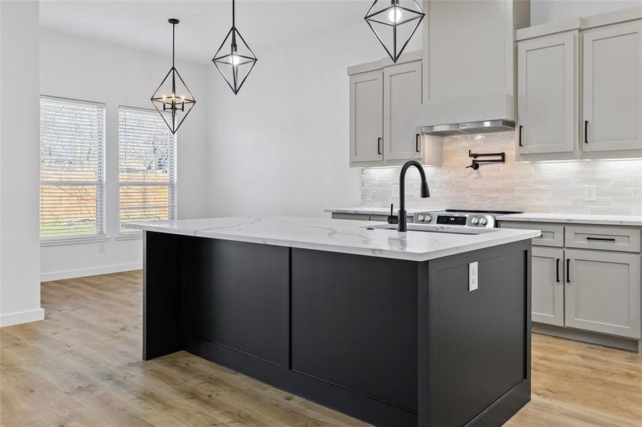 Kitchen with light wood finished floors, an island with sink, light stone counters, backsplash, and wall chimney range hood