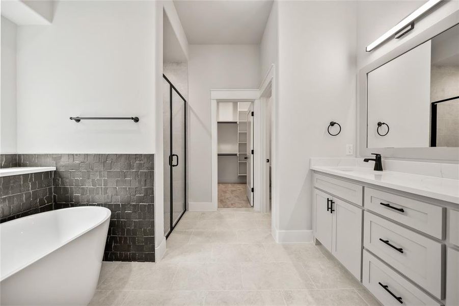 Bathroom featuring a freestanding tub, dark tile wainscoting, glass-enclosed shower, single vanity with quartz countertop, and light tile flooring