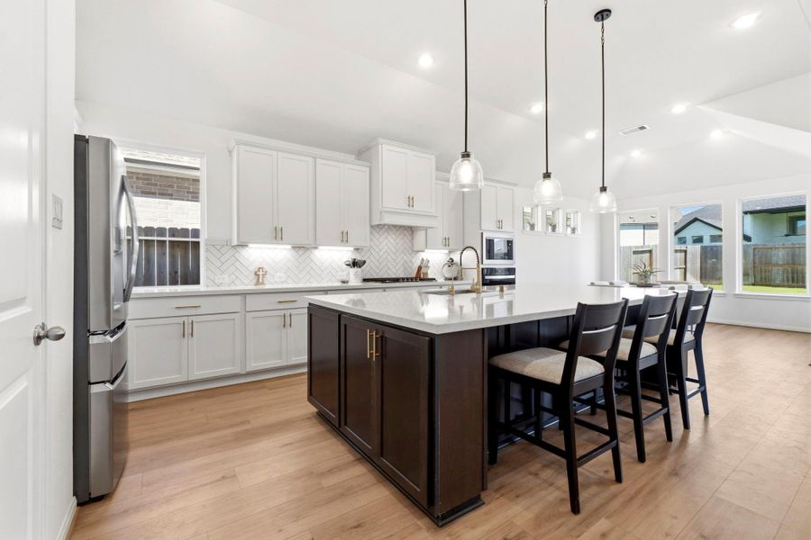 This modern kitchen features a large island with room for seating, sleek pendant lighting, stainless steel appliances, and white cabinetry with a herringbone backsplash. Ample natural light fills the space through multiple windows.