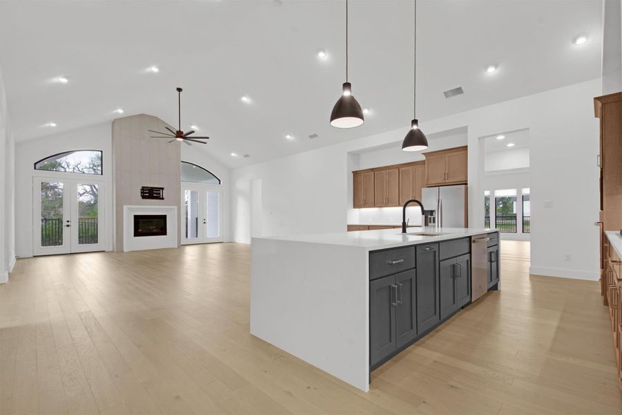 Kitchen featuring 16ft vaulted ceiling, generous amount of natural light, a spacious island with pendant lighting, quartz countertops, light hardwood floors, gas fireplace, and double french doors to outdoor patio with scenic, greenbelt view