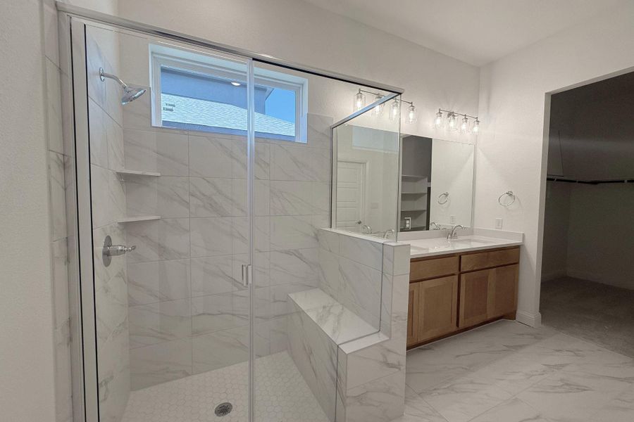 Spacious bathroom featuring a glass-enclosed shower with a built-in bench, dual vanity with a light wood cabinet, large format tiled flooring, and a window for natural illumination