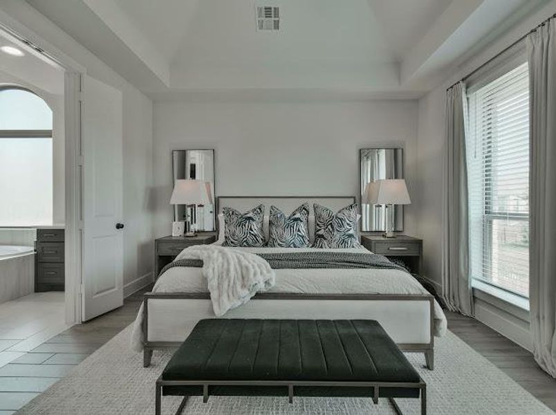 Bedroom featuring multiple windows, light wood-type flooring, connected bathroom, a raised ceiling, and vaulted ceiling Bedroom featuring multiple windows, light wood-type flooring, connected bathroom, a raised ceiling, and vaulted ceiling