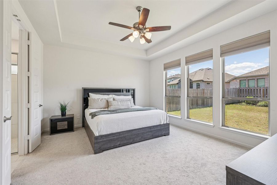 This room features a tray ceiling with a ceiling fan, three large windows overlooking the backyard, and light-colored carpeting