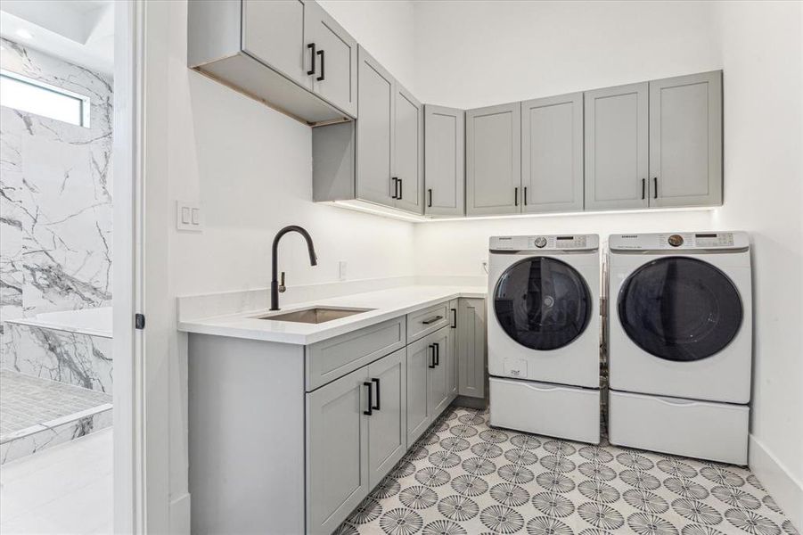 This utility room features built-in cabinetry for storage, a sleek countertop with an integrated sink, patterned tile flooring, and space for a washer and dryer, offering both functionality and style.