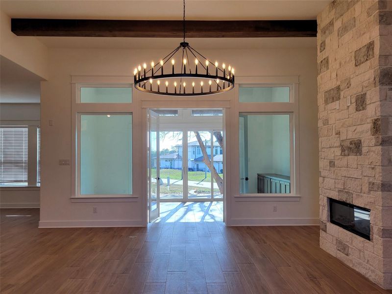 This room features a modern, open-concept design with a large window and glass doors offering ample natural light. A striking stone fireplace adds warmth, while a stylish chandelier and wood beam ceiling enhance the space's elegance. This room features a modern, open-concept design with a large window and glass doors offering ample natural light. A striking stone fireplace adds warmth, while a stylish chandelier and wood beam ceiling enhance the space's elegance.