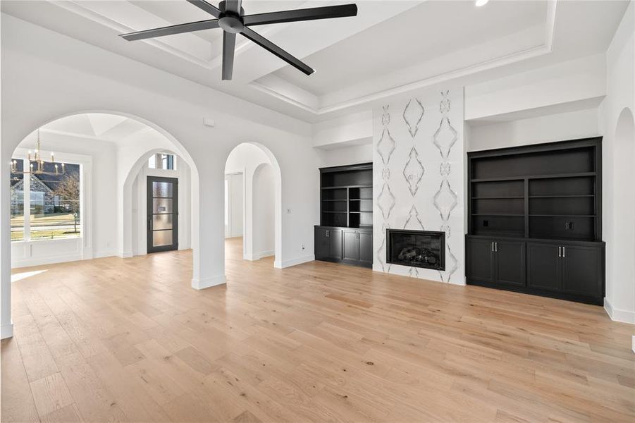 Unfurnished living room with arched walkways, light wood-style floors, built in features, a tray ceiling, and a premium fireplace Unfurnished living room with arched walkways, light wood-style floors, built in features, a tray ceiling, and a premium fireplace