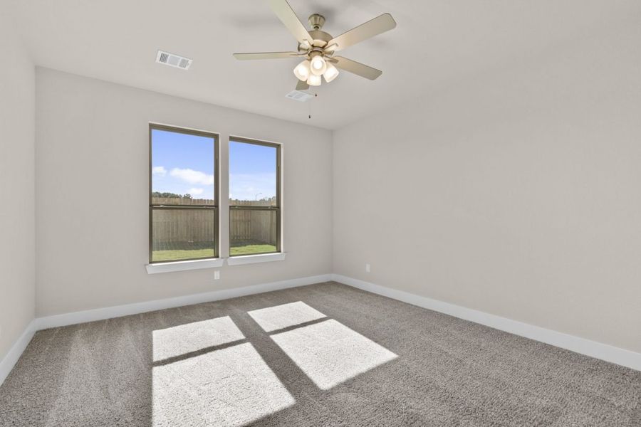 Image of a bedroom with beige walls, tan carpeting, windows with black trim, and a ceiling fan