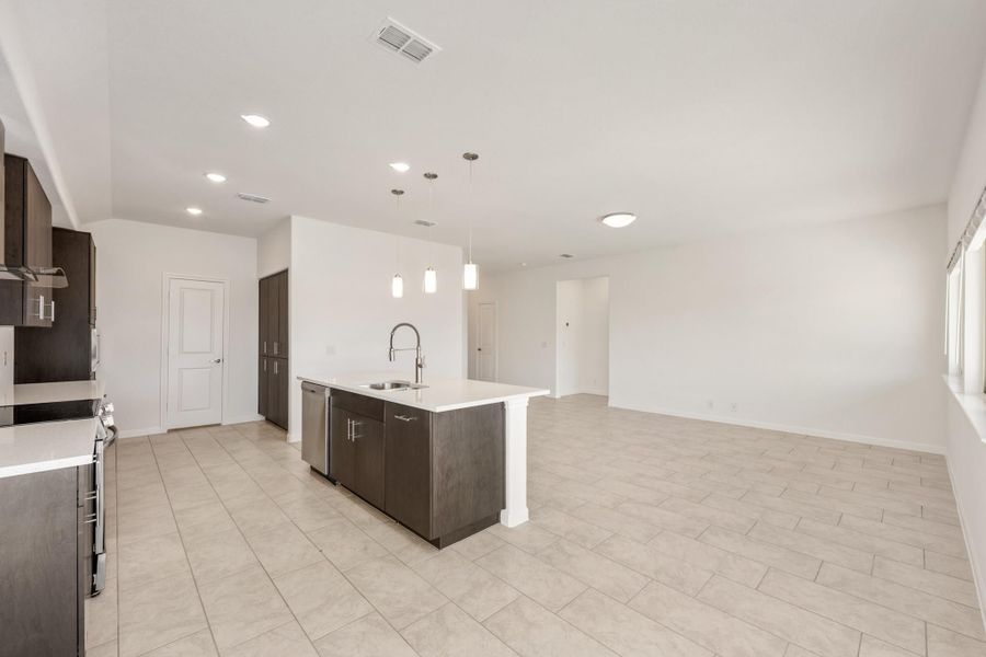 Open kitchen with dark cabinets, center island with sink, pendant lights, and tile floors opening to living area
