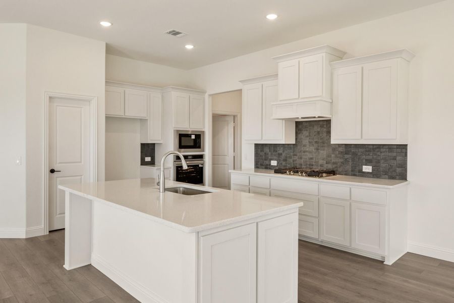 Kitchen with white cabinets, large island with sink, gray tile backsplash, and stainless steel appliances on wood-look floors. Kitchen with white cabinets, large island with sink, gray tile backsplash, and stainless steel appliances on wood-look floors.