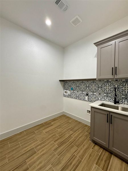 This photo shows a modern laundry room with stylish patterned backsplash, light gray cabinetry, and wood-look tile flooring. The space is bright and functional, featuring a sink and washer/dryer hookups. This photo shows a modern laundry room with stylish patterned backsplash, light gray cabinetry, and wood-look tile flooring. The space is bright and functional, featuring a sink and washer/dryer hookups.