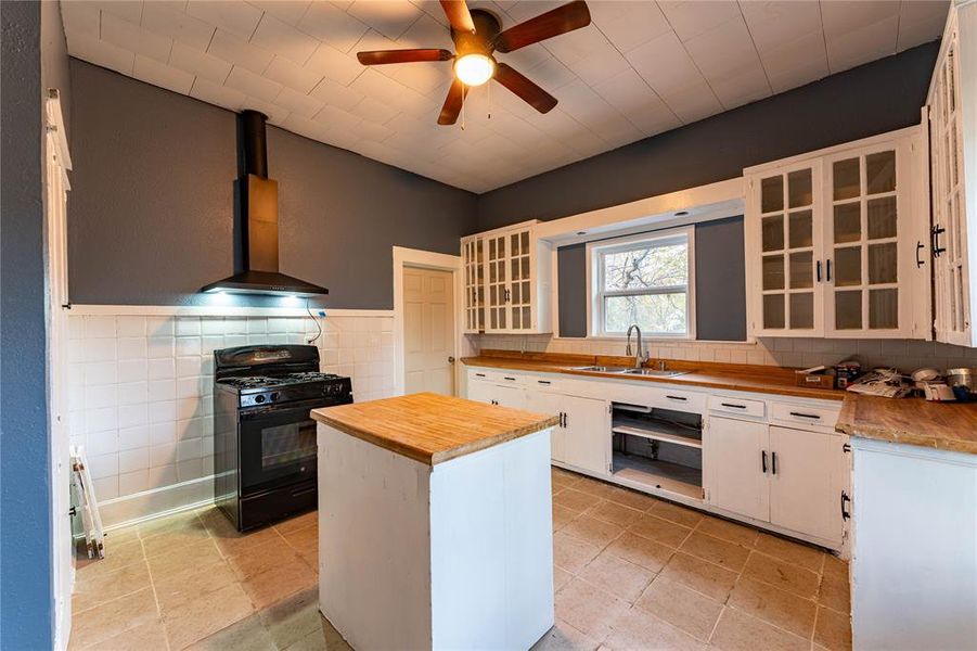 Kitchen featuring wood counters, wall chimney exhaust hood, sink, white cabinets, and black gas range Kitchen featuring wood counters, wall chimney exhaust hood, sink, white cabinets, and black gas range