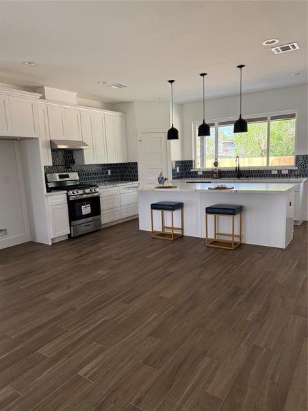 This photo showcases a modern kitchen with white cabinetry, stainless steel appliances, a central island with bar stool seating, dark mosaic backsplash, and rich, wood-toned flooring. The space is well-lit with natural light and elegant pendant lighting.