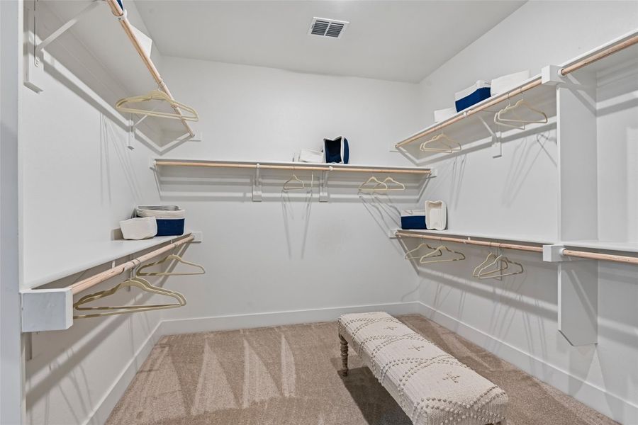 Walk-in closet with white shelving, multiple hanging rods, carpet flooring, and a small upholstered bench Walk-in closet with white shelving, multiple hanging rods, carpet flooring, and a small upholstered bench