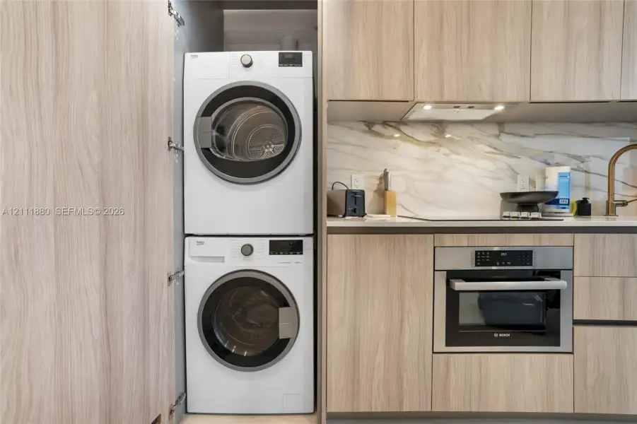 Stacked washer and dryer unit, wood-finish cabinetry, built-in stainless steel oven, and stone-surface backsplash