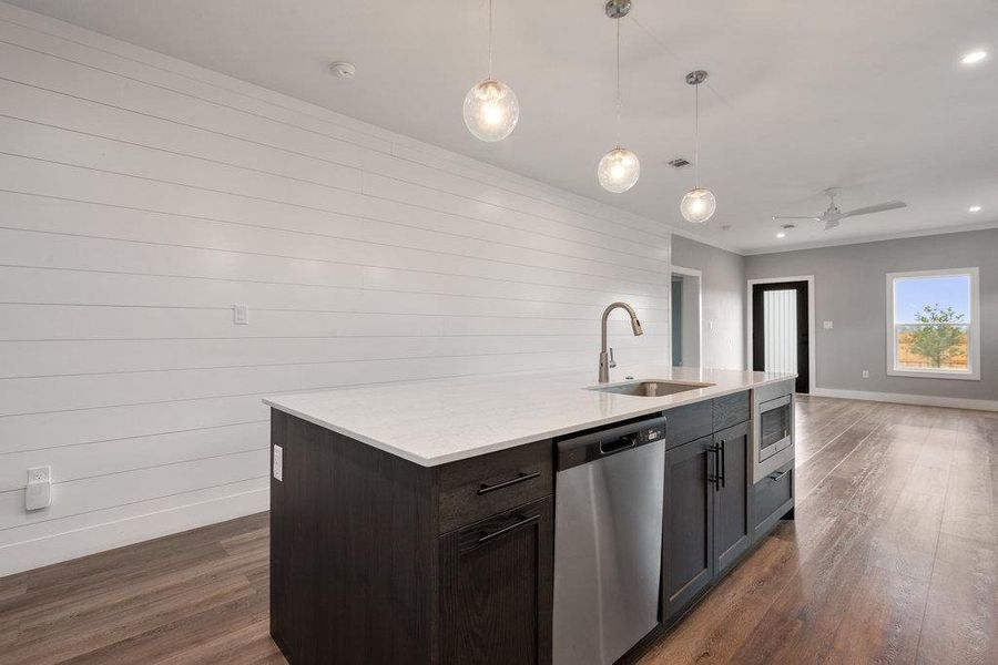 Kitchen featuring stainless steel appliances, hanging light fixtures, dark wood-style floors, a center island with sink, and light stone counters