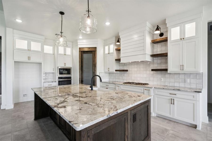 Kitchen featuring premium range hood, decorative light fixtures, backsplash, a kitchen island with sink, and white cabinetry