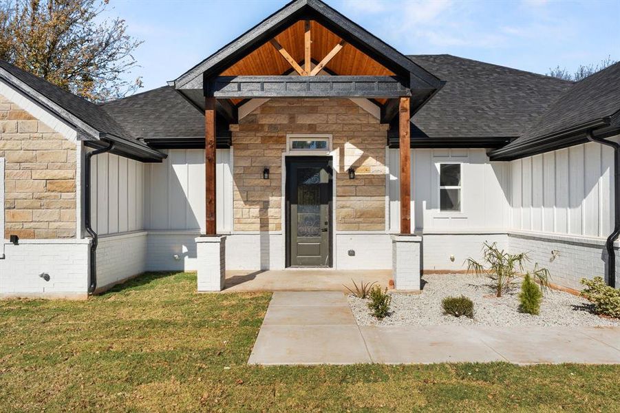 Doorway to property with board and batten siding, roof with shingles, a lawn, and covered porch