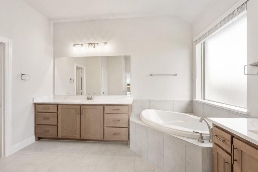 Master bathroom with wood vanity, large mirror, soaking tub, and tile surround near a bright window