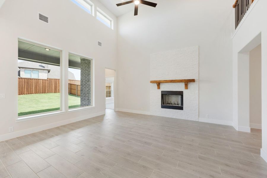 Open living room with two-story ceiling, white brick fireplace, wood mantel, and large windows overlooking backyard