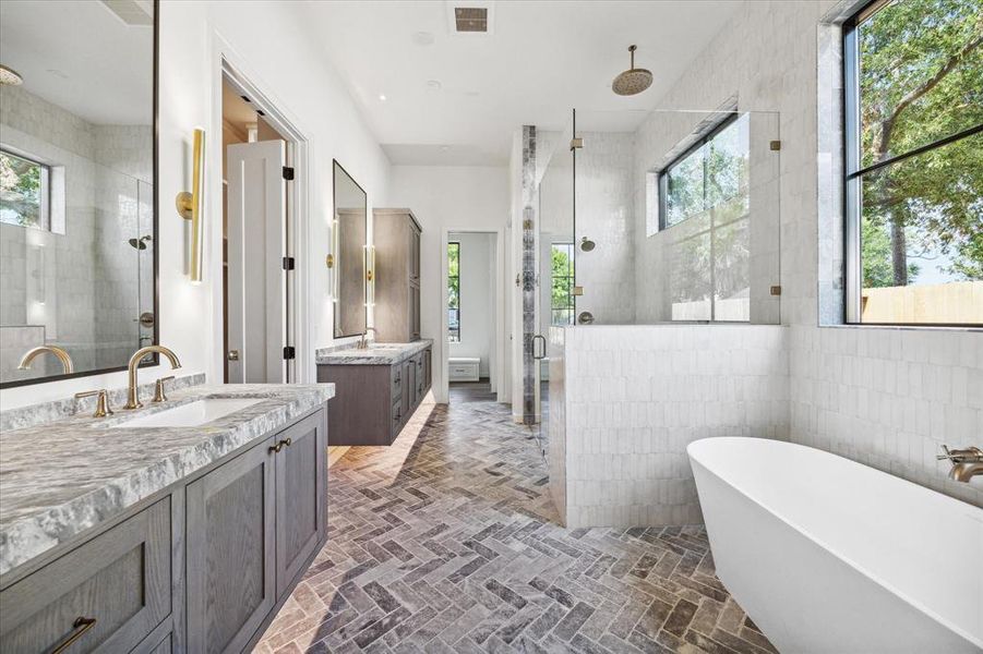 This luxurious primary bathroom features a double vanity with elegant marble countertops, a spacious walk-in shower with seamless glass and dual shower heads, and is complemented by a freestanding soaking tub. Large windows fill the space with natural light, highlighting the intricate herringbone tile flooring and the overall sophisticated design. The tile surround goes all the way to the ceiling for the complete package luxury feel for the dual walk-in closets.