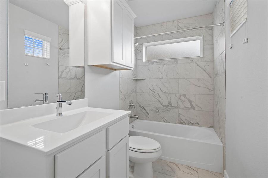 Secondary bath features tile flooring, bath/shower combo with tile surround, white stained wood cabinets, beautiful light countertops, mirror, dark, sleek fixtures and modern finishes.