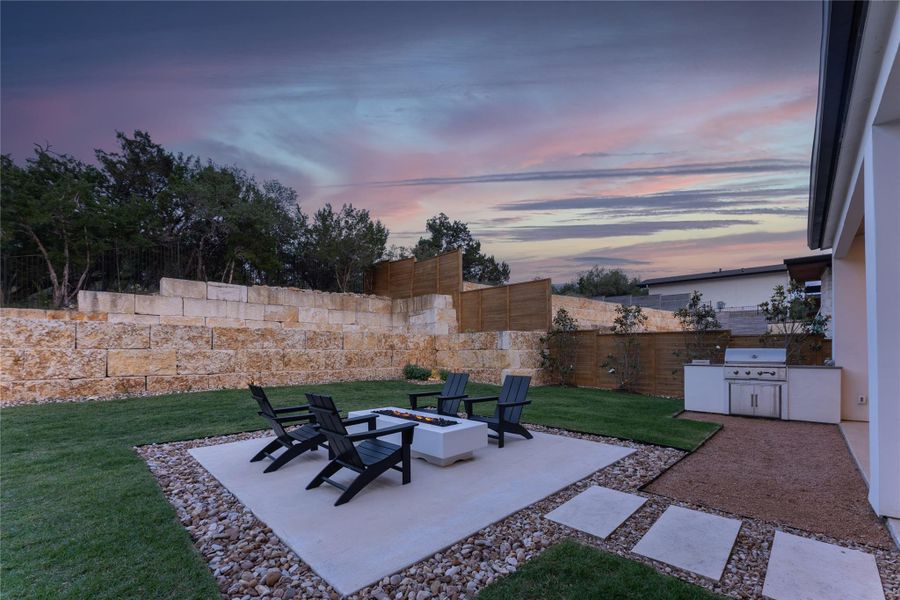 Expansive backyard featuring a built-in outdoor kitchen with grill, a fire pit area with concrete patio, a tiered stone retaining wall, mature perimeter trees, and stepping stone path Expansive backyard featuring a built-in outdoor kitchen with grill, a fire pit area with concrete patio, a tiered stone retaining wall, mature perimeter trees, and stepping stone path