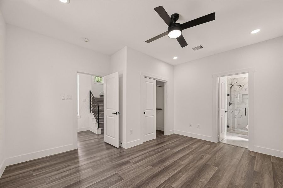 This photo shows a modern, spacious room with light gray walls and dark wood flooring. It features a ceiling fan, recessed lighting, and a view of a staircase. There are two doors, one leading to a walk-in closet and another to a bathroom with marble accents. This photo shows a modern, spacious room with light gray walls and dark wood flooring. It features a ceiling fan, recessed lighting, and a view of a staircase. There are two doors, one leading to a walk-in closet and another to a bathroom with marble accents.