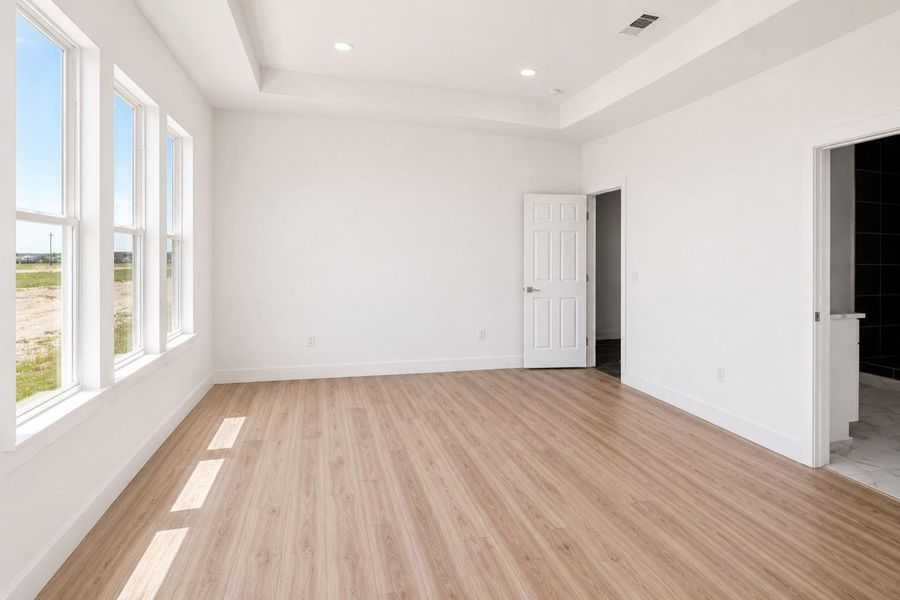 A generously sized primary bedroom with light wood flooring and tray ceilings offers ample natural light.