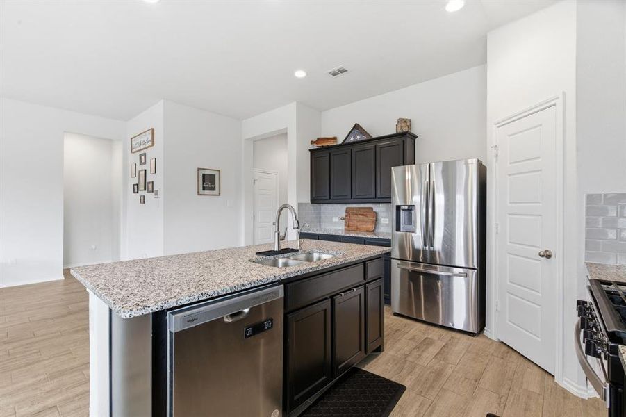 Kitchen with appliances with stainless steel finishes, backsplash, a center island with sink, light stone countertops, and light wood-style flooring