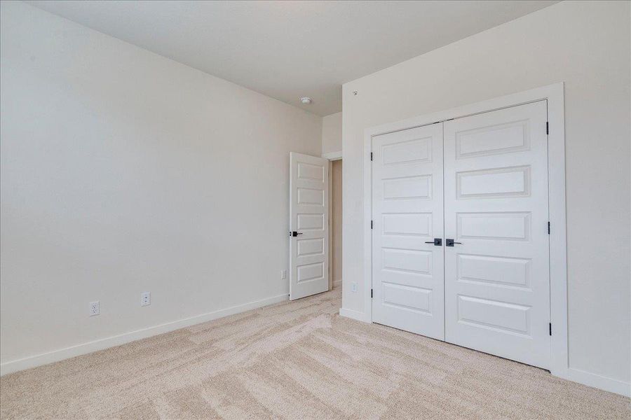 A well-lit bedroom features plush carpet flooring and clean, paneled doors.