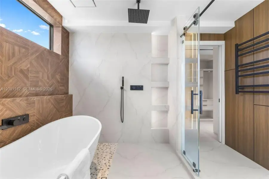 Freestanding soaking tub with matte black wall-mounted faucet, walk-in shower with glass sliding door, rainfall showerhead, integrated tiled shelving, and wood-finish wall accents Freestanding soaking tub with matte black wall-mounted faucet, walk-in shower with glass sliding door, rainfall showerhead, integrated tiled shelving, and wood-finish wall accents