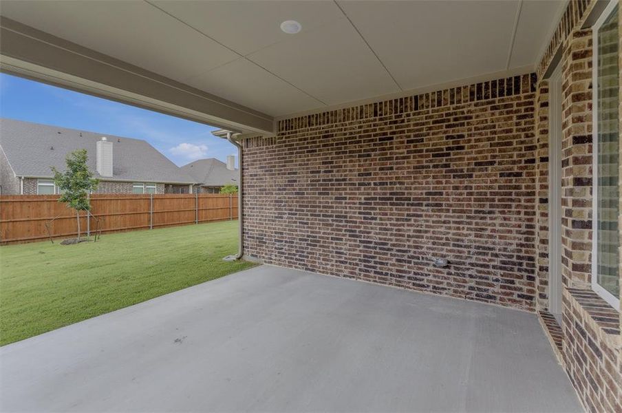 Covered concrete patio featuring a brick exterior wall, recessed lighting, and a view of the fenced backyard with green lawn