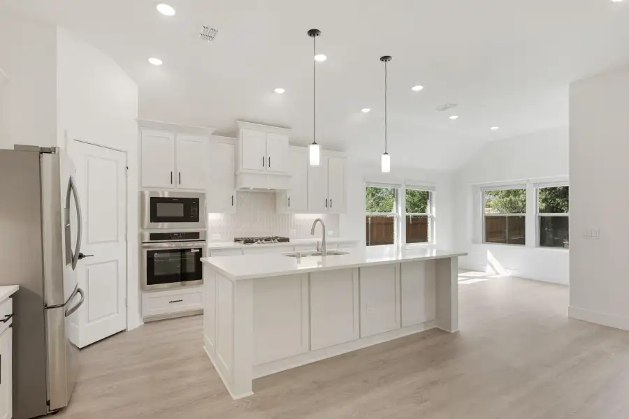 White kitchen with large island, stainless steel appliances, pendant lights, and light wood floors open to living area