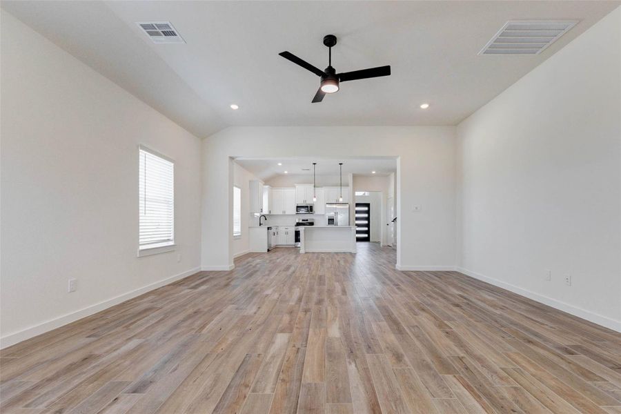 This expansive open-concept living area showcases a seamless flow into the designer kitchen, creating the perfect space for entertaining and everyday living. Featuring rich wood-look tile flooring, soaring ceilings, and a modern ceiling fan, the room feels both spacious and inviting. Natural light pours in through multiple windows, while the wide opening frames the bright white kitchen, complete with an oversized island and stylish finishes.