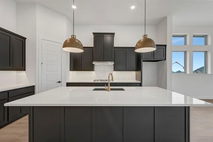 Kitchen with light wood-style floors, an island with sink, light stone counters, and decorative light fixtures