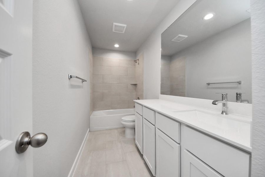 Secondary bath features tile flooring, bath/shower combo with tile surround, light stained wood cabinets, beautiful light countertop.