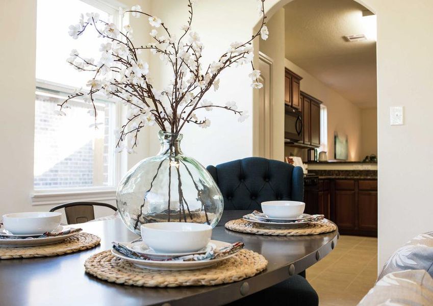 Eating area with glass vase that has decorative stick plants, place setting with white bowl, both sitting on dark brown wooden table