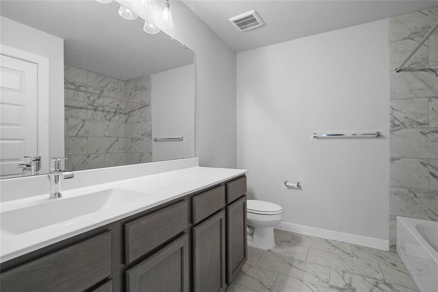 Secondary bath features tile flooring, bath/shower combo with tile surround, stained wood cabinets, beautiful light countertops, mirror, dark, sleek fixtures and modern finishes.