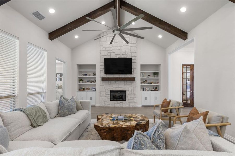 Living room with a stone fireplace, wood finished floors, beamed ceiling, high vaulted ceiling, and ceiling fan Living room with a stone fireplace, wood finished floors, beamed ceiling, high vaulted ceiling, and ceiling fan