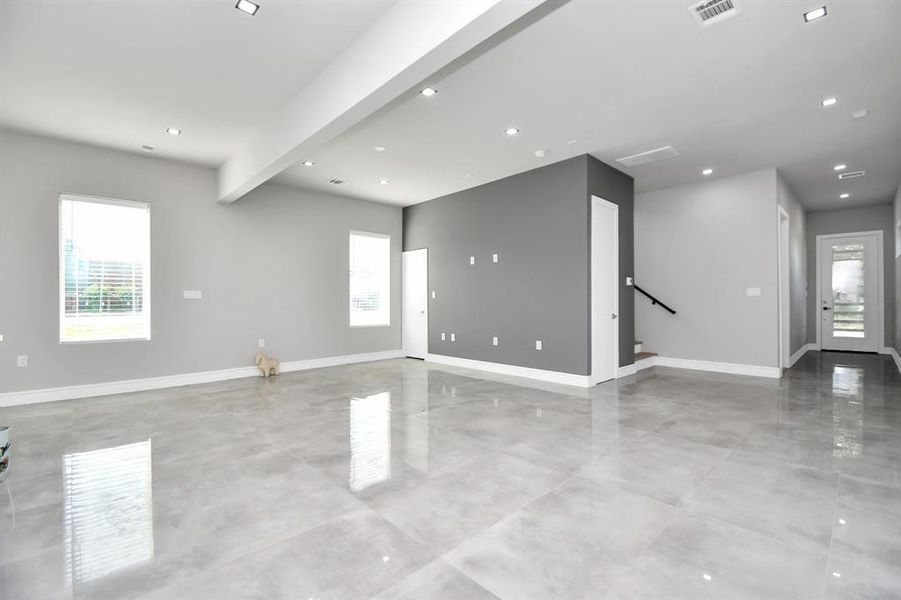 This is a modern, open-plan living space with sleek finishes, featuring a large kitchen island, ample natural light from several windows, and recessed lighting. The room presents a clean, minimalist aesthetic with neutral colors.