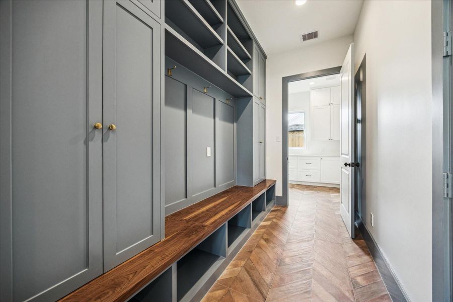 Custom mud room showcases full height cabinetry, open cubbies, and a rich wood bench that adds warmth against the slate blue millwork. Herringbone tile flooring and brass hooks create a polished drop zone just off the garage entry.