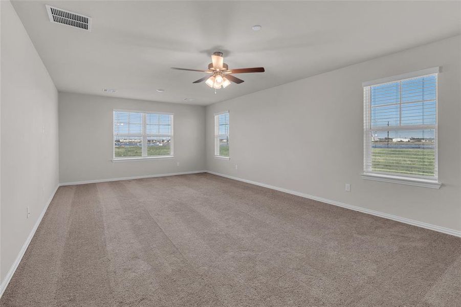 This is a spacious Primary bedroom, well-lit with neutral walls and carpet flooring. It features a ceiling fan with lighting and two large windows that provide ample natural light and a view of the outside.