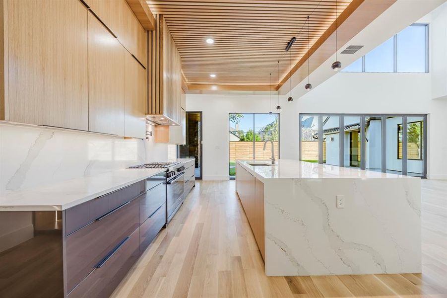 Kitchen with modern cabinets, light stone counters, light wood finished floors, a large island with sink, and stainless steel range with gas cooktop Kitchen with modern cabinets, light stone counters, light wood finished floors, a large island with sink, and stainless steel range with gas cooktop