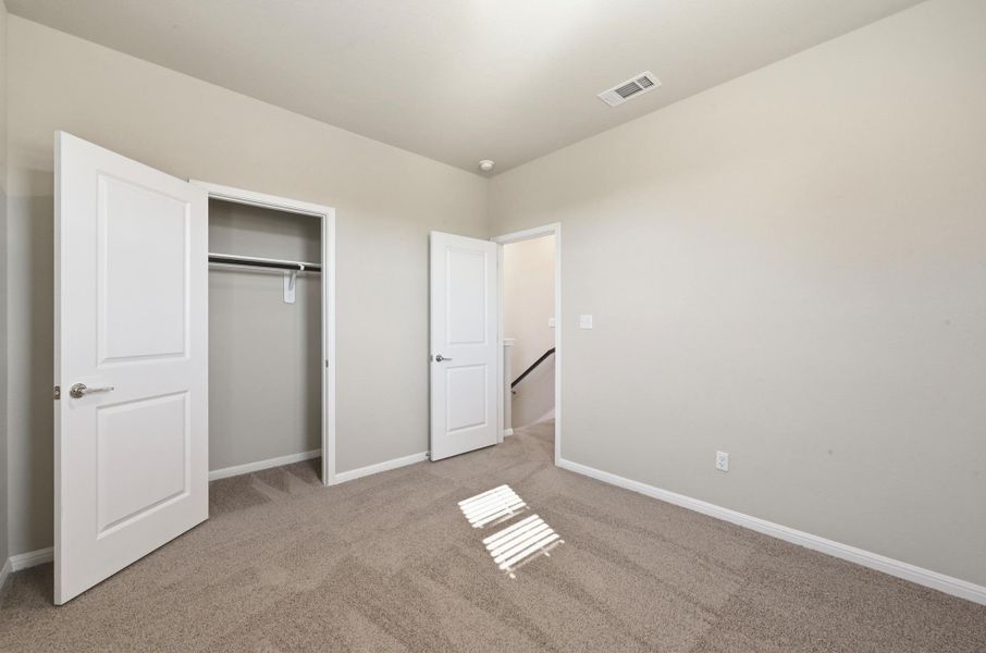 Neutral-toned bedroom featuring plush carpet flooring, a built-in closet with double panel doors, and a staircase visible through an open doorway Neutral-toned bedroom featuring plush carpet flooring, a built-in closet with double panel doors, and a staircase visible through an open doorway