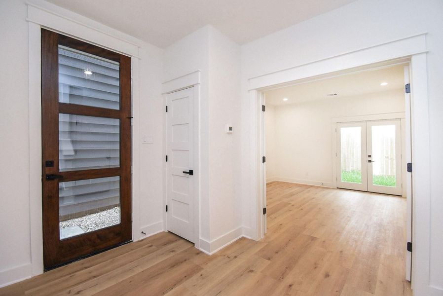 This photo showcases a bright entryway with light wood flooring and a modern wood-framed glass door. Adjacent is a spacious room with French doors leading to a small outdoor area, enhancing the open and airy feel.