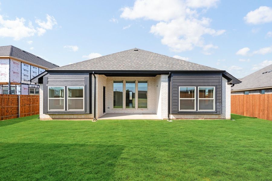 Rear exterior of single-story home with brick and dark siding, large windows, patio, and fenced green backyard