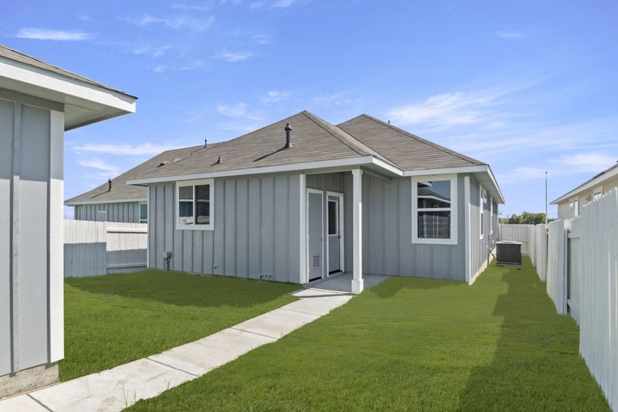 Image of exterior rear of a one-story grey-blue home with sidewalk to detached garage, green grass, white fence, and sky